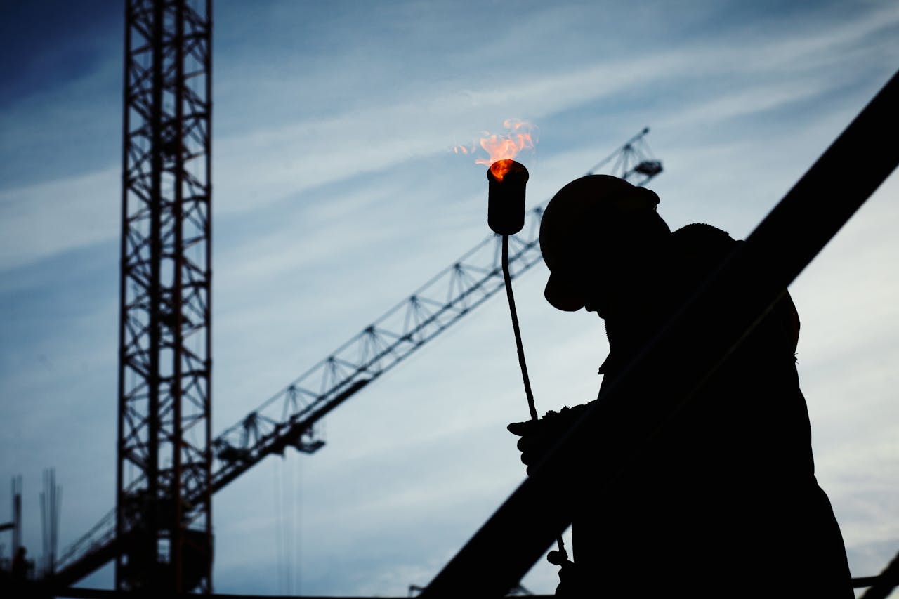 contact-img Silhouette of a construction worker using a blowtorch at a building site against a crane-filled skyline.